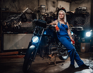 Tattooed hipster girl in work overalls hold a big wrench and posing for a camera while leaning on her naked bike in garage or workshop
