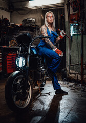Tattooed female mechanic in work overalls hold a big wrench and posing for a camera while leaning on her naked bike in garage or workshop