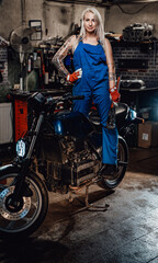 Plakat Tattooed hipster girl in work overalls hold a big wrench while standing on naked bike in garage or workshop, smiling and looking on camera