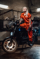 Naklejka na ściany i meble Tattooed hipster girl in orange overalls hold a big wrench and posing for a camera while standing on her naked bike in garage or workshop