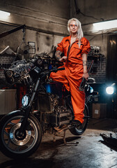 Fototapeta na wymiar Tattooed hipster girl in orange overalls hold a big wrench and posing for a camera while standing on her naked bike in garage or workshop, smiling and looking on camera