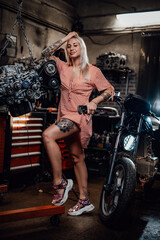 Fototapeta na wymiar Beautiful blonde girl with tattooed body wearing pink dress posing next to the suspended flat engine and naked bike in garage or workshop, smiling and looking on camera