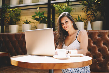 Portrait of young attractive woman connect via wireless to internet while she sitting in modern coffee shop interior,female freelancer working on laptop computer while she breakfast with cake and cafe