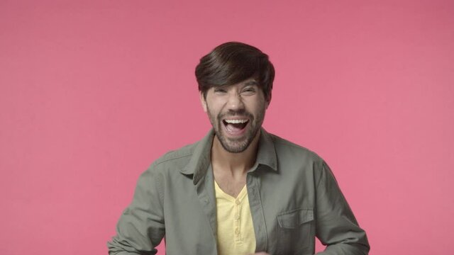 Handsome bearded adult man look camera starts laughing over hilarious tv series, joking, chuckle and clap hands as watching stand-up comedy performance, standing pink background