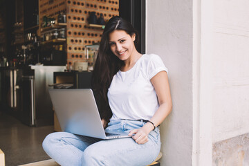 Smiling latin woman looking at you while working on net-book during lunch break, charming female freelancer using laptop computer for remote job while sitting in modern coffee chop outdoors