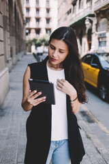 Portrait of a young female student reading news on touch pad while standing in urban setting, modern tourist woman dressed in stylish clothes using digital tablet computer while walking on the street
