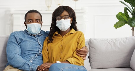 Portrait African American Couple Sitting On Sofa In Protective Medical Masks.