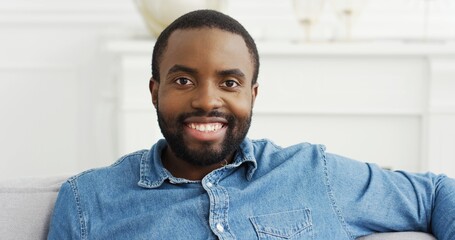 Close up portrait of happy bearded African American man while sitting on sofa at his modern home.