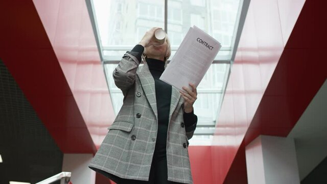 A bottom view slow motion close up video of a blonde female executive drinking coffee from a plastic cup and reading a recieved contract