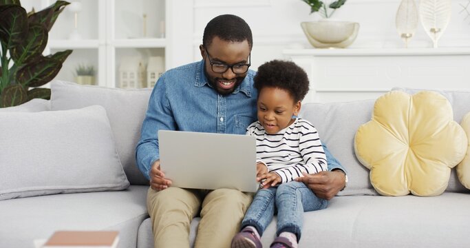 Portrait of african american father sitting with daughter on sofa using laptop watching video.