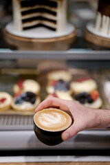 Barista holds a ready-made latte of coffee in a black mug against the backdrop of a chicken with sweet desserts in a pastry shop.