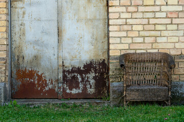 An old wicker chair stands outside near a brick house. Free seat on the armchair. A cozy place to relax in nature.