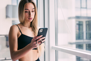 Smiling young woman using phone while sitting near window in living room