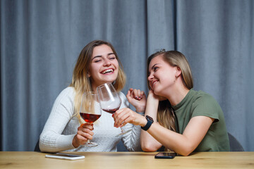 two girls of European appearance with blond hair are sitting at the table, drinking wine and laughing, relaxing at home, alcohol