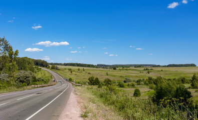 A beautiful asphalt road passes through a blooming green field and forest