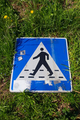 Broken spent blue cross walk sign laying on green grass.