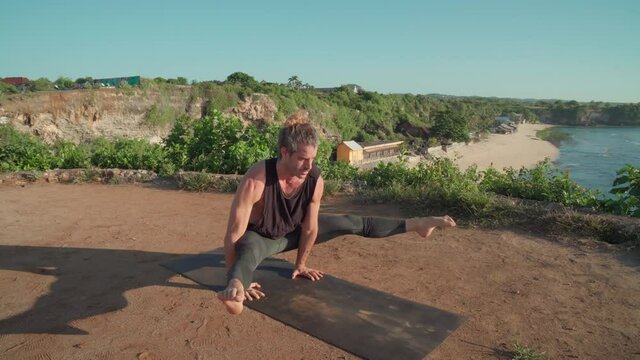 Tracking shot of muscled male yogi doing ashtavakrasana, straddle press handstand and stretching on mat while practicing on cliff overlooking ocean on coast of Bali