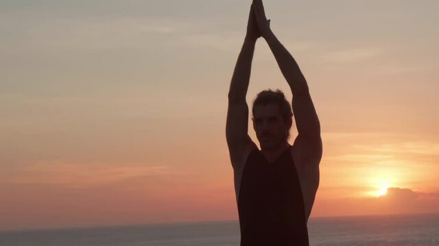 Handheld tracking shot of muscly male yogi standing in tree pose on cliff by ocean at sunset