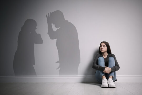 Upset teenage girl sitting on floor near wall and silhouettes of arguing parents