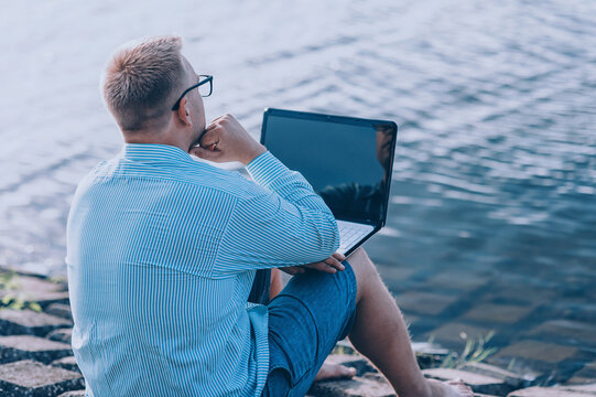 A man in glasses and a shirt working on a laptop in the fresh air, against the background of a river.