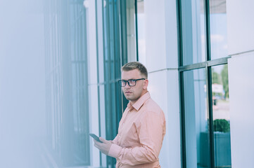 Businessman in a pink shirt with a smartphone in his hand stands against the backdrop of a modern building with large windows.