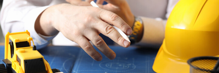 Man holds pencil, on table yellow helmet and truck. Construction calculation for use rented...
