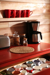 Open drawer with cups and coffeemaker near wooden wall indoors