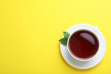 Cup of aromatic black tea and green leaves on yellow background, top view. Space for text
