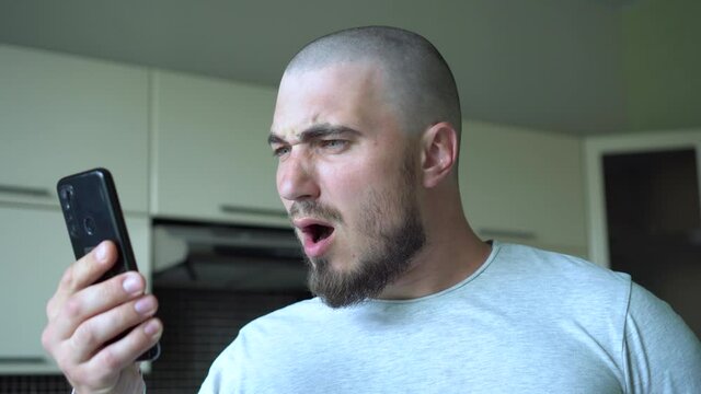 A Bald and Attractive Man with a Beard is Shocked by the News. Reads News on Smartphone. Stress, Dumbfounded, Shock. Worried young man reading bad news in cell phone at home.