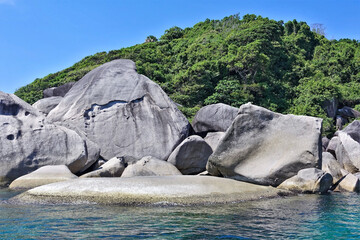 Amazing Similan island. On the shores of the turquoise sea lie huge ancient smooth boulders. Behind them rises a hill overgrown with dense tropical forest. Bright blue sky, sunny day. Thailand.