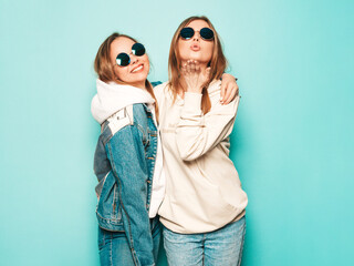 Two young beautiful brunette smiling hipster girls in trendy summer hoodie and jeans jacket clothes. Sexy carefree women posing near blue wall. Trendy and positive models give air kiss in sunglasses