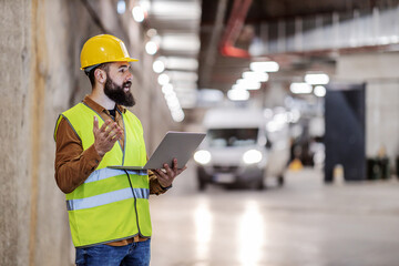 Young serious attractive contractor standing with laptop in hands and giving orders to workers.