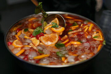 Fruit cocktail in a large metal pan with a ladle. Apples, lemons, mint, oranges, strawberries with red wine and juice, with ice. A party. Selective focus