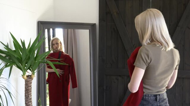 blonde girl chooses is dressing up a red coat for autumn season at mirror at home