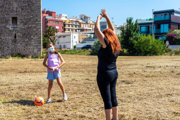 Mother and daughter playing soccer with an orange ball in a field with grass wearing masks to prevent coronavirus