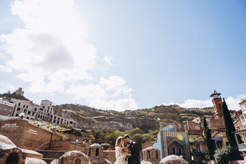Stylish couple of brides: Georgian-style wedding, man with mustache dressed in costume and pretty blonde bride in wedding dress against beautiful city background