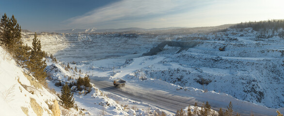 Panorama of a large stone quarry covered with deep snow on a frosty winter day with a heavy mining truck in the foreground. Mining industry.
