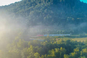 Aerial drone view of a passenger train travelling at early morning in a valley ful of misty fog