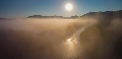 Aerial drone panorama of river and fog over misty plains with hills perking out of the fog during early sunrise hours. Early morning on river and fields.