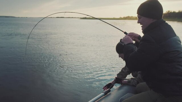 Friends fishing for trophy. Two friends fishing from the boat on the calm lake during sunset. Man holds the bending rod and fights with big catfish