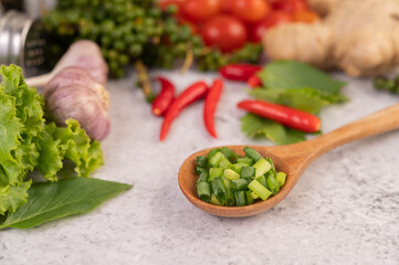 Chopped spring onion on a wooden spoon with peppers and tomatoes on a white cement floor.