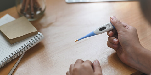 A hand is holding a digital thermometer over the wooden working desk surrounded by a notebook and pencil holder.