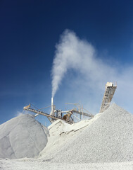 High column of dust over the stone crushing equipment at the mining enterprise on the background of the blue sky, vertical panorama. Mining industry.