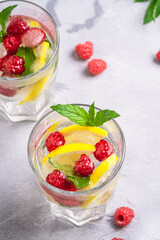 Fresh cold ice water drink with lemon, raspberry fruits and mint leaf in two faceted glass on stone concrete background, summer diet beverage, angle view selective focus
