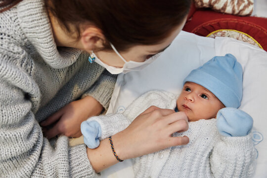 Young Redheaded Mother With Preventive Health Mask And Lgtbiq+ Bracelet, Next To Her Newborn Baby Playing And Laughing Together On The Bed