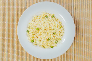 Cooked jasmine rice with green peas on a white plate and bamboo table surface. Top down view,