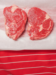 Two raw premium rib eye steaks on white paper and professional red and white stripes butcher apron.