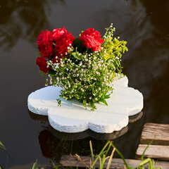 A square photo of a bouquet of beautiful roses on the water which reflects the sky and trees and greenery in the foreground