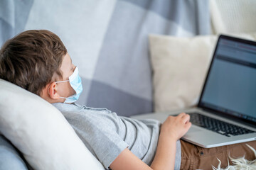 Focused boy in a mask, lying on couch with laptop during the quarantine period.