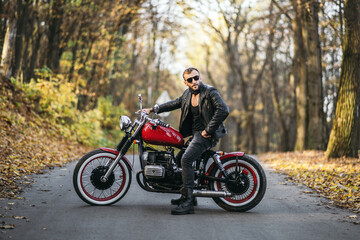 Naklejka na ściany i meble Bearded brutal man in sunglasses and leather jacket sitting on a motorcycle on the road in the forest